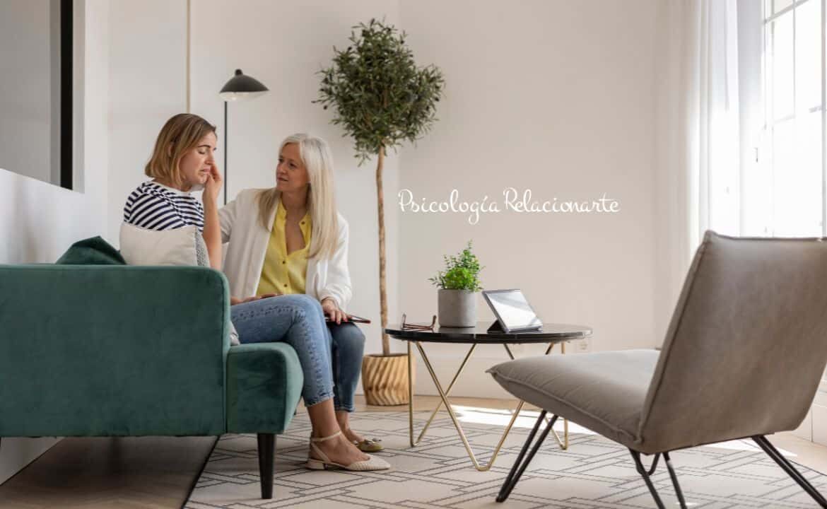 Acompañar el estrés postraumático en una consulta psicológica: dos mujeres sentadas en un sofá mientras una ofrece apoyo y escucha empática a la otra.