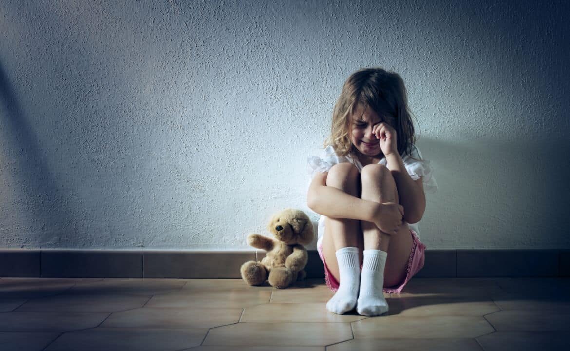 Niña sentada en el suelo junto a osito de peluche, llorando víctima de maltrato infantil.