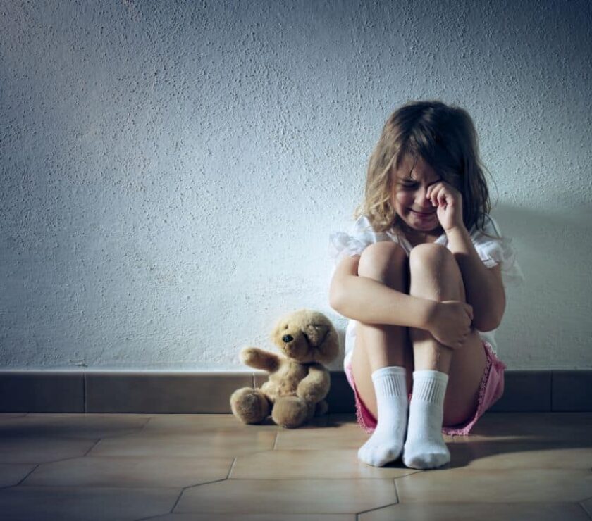 Niña sentada en el suelo junto a osito de peluche, llorando víctima de maltrato infantil.