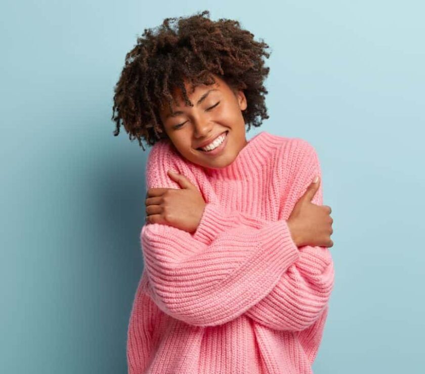 Mujer joven sonriente con jersey de lana rosa abrazándose con autoestima saludable