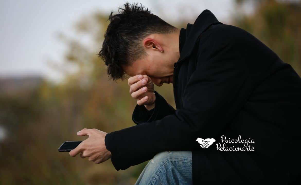 Joven sentado con abrigo negro y móvil en la mano, sufriendo intentando aliviar el dolor emocional
