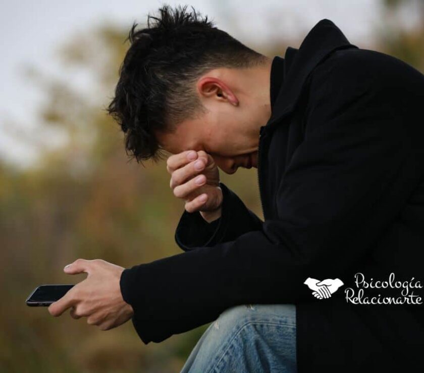 Joven sentado con abrigo negro y móvil en la mano, sufriendo intentando aliviar el dolor emocional