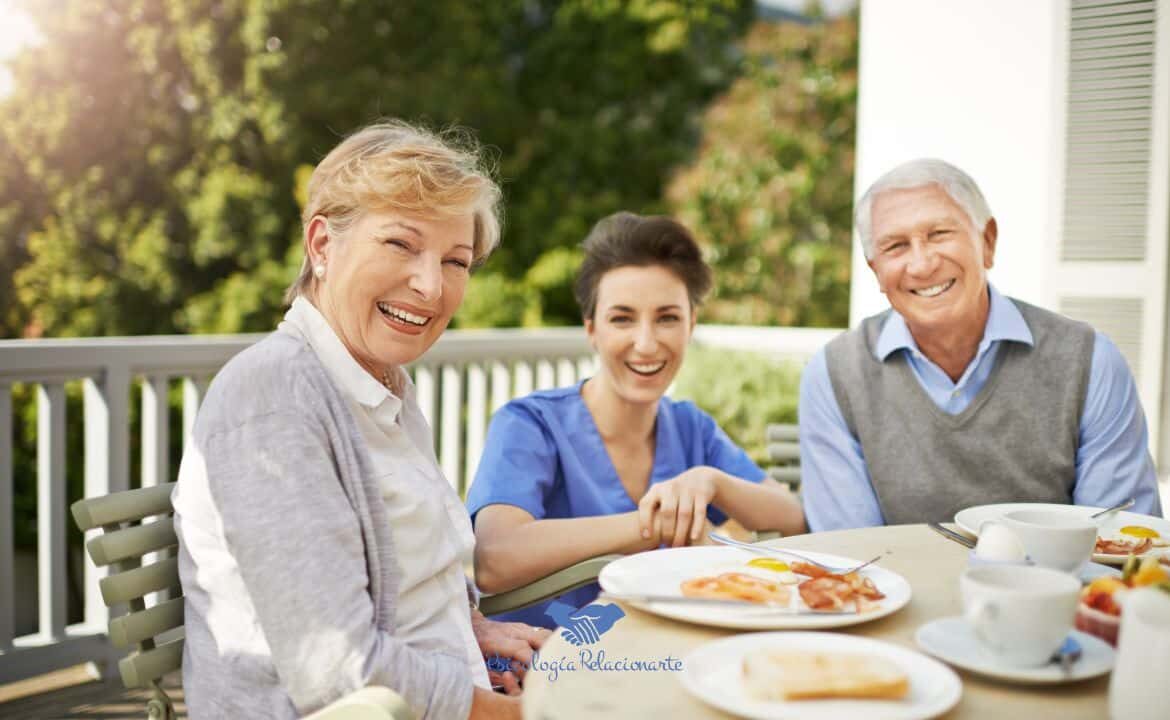 Una pareja senior y cuidadora sentados a la mesa compartiendo felicidad y calidad de vida.