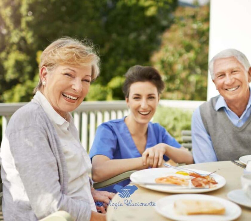 Una pareja senior y cuidadora sentados a la mesa compartiendo felicidad y calidad de vida.